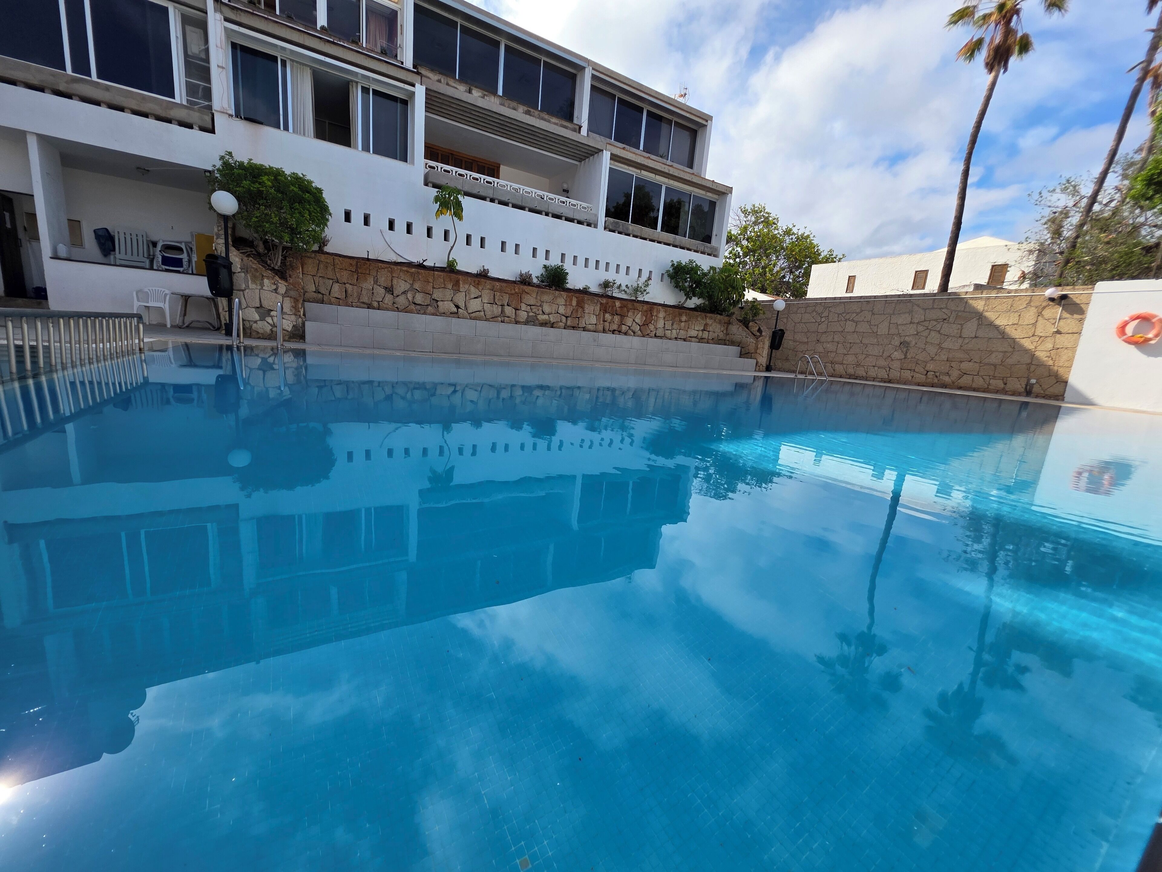 Une piscine à l’eau d’un bleu cristallin, entourée d’un mur de pierre et bordée d’un bâtiment blanc doté de balcons.