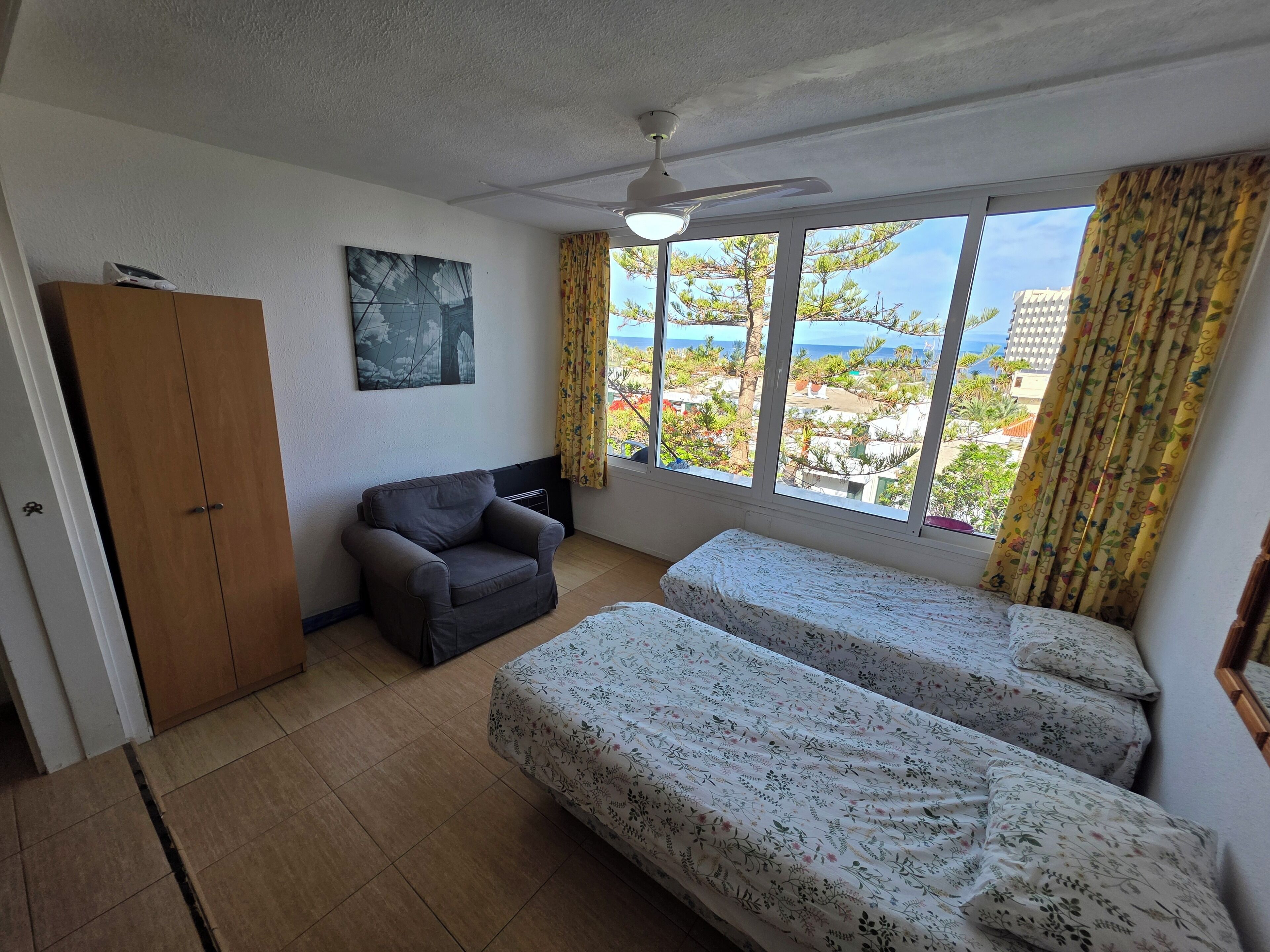 Una habitación con cama, una silla, un armario, un ventilador de techo y una ventana con cortinas.