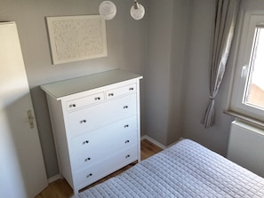 A bedroom with a white dresser, a bed with a quilted cover, a window with curtains, and a wall-mounted light fixture. - Maisonette charming apartment, near bike and. Hiking trails in the Nassau / Limburg nature park (Altendiez)