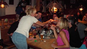 Een groep mensen proostend aan een houten tafel in een donkere bar, met hangende lampen en houten balken.