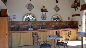 An outdoor kitchen area with a brick backsplash, two arched windows, and a rustic wooden table and chairs.