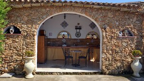 A stone-walled outdoor seating area with a tiled roof, two chairs, and a table.