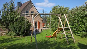 A wooden swing set and slide in a grassy yard with a brick house in the background. - A lot of space and rest between beach and nature (Sagard)