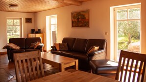 A cozy living room with a wooden ceiling, a brown leather sofa, a wooden coffee table, and a view of the outdoors through large windows. - A lot of space and rest between beach and nature (Sagard)