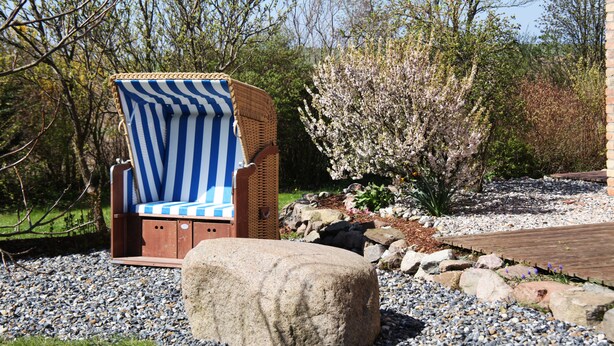 A wicker chair with blue and white stripes, a stone bench, and a garden with rocks and flowering plants. - A lot of space and rest between beach and nature (Sagard)
