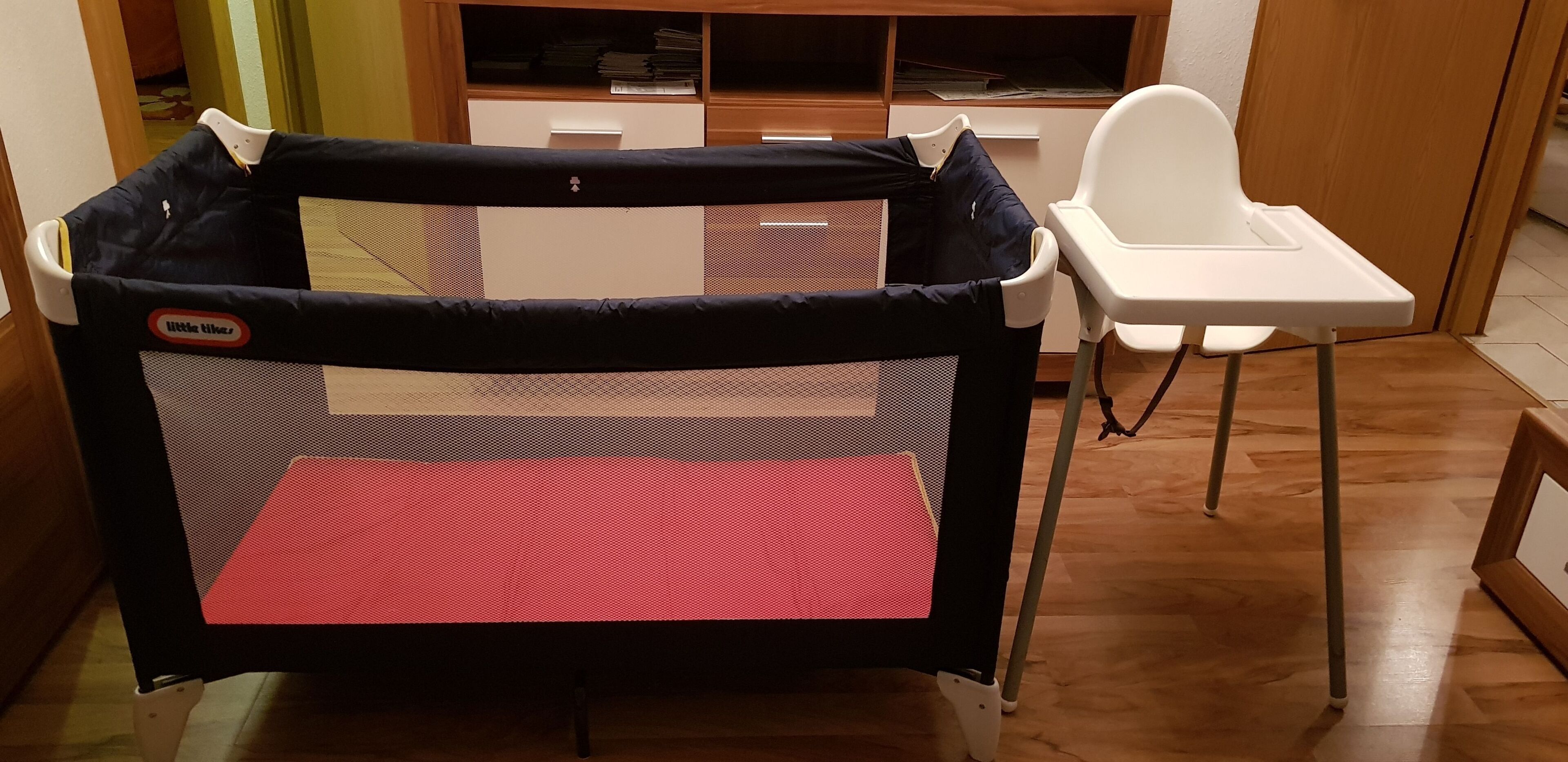 A room with a baby playpen, a white high chair, and a wooden floor.