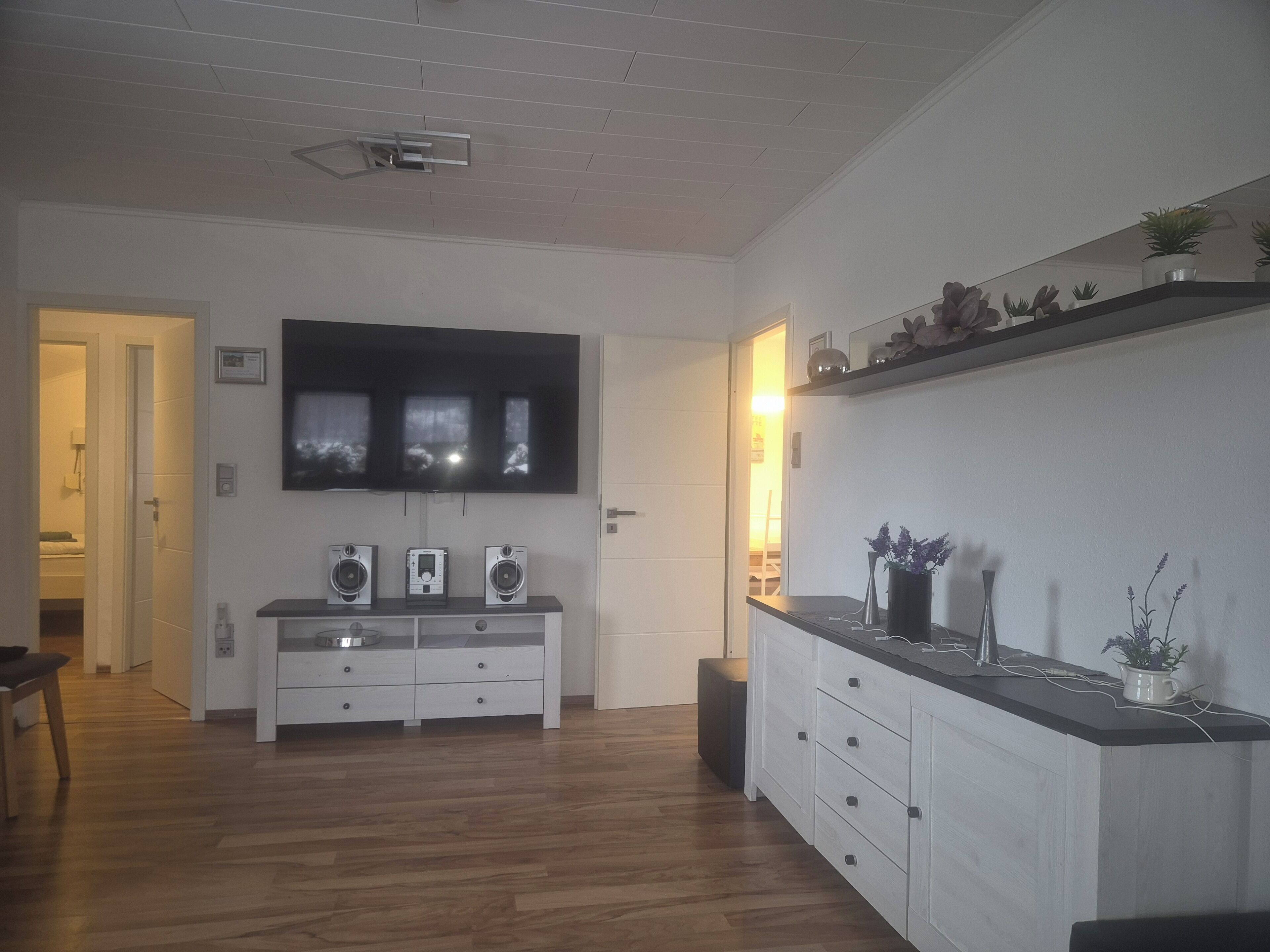 Un salon moderne avec un téléviseur à écran plat fixé au mur, un meuble de rangement blanc avec des haut-parleurs intégrés et un plancher en bois.