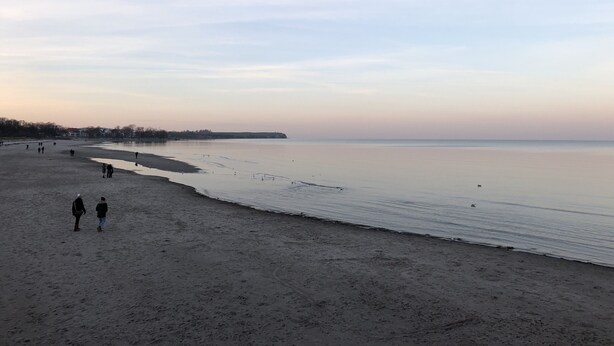 Ein Strand mit ruhiger See, Menschen, die am Ufer entlanggehen, und eine entfernte Küstenlinie mit Gebäuden.