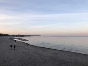 Une plage avec une mer calme, des gens qui se promènent le long du rivage et une ligne côtière lointaine parsemée de bâtiments.