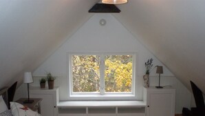 Une chambre avec un plafond en pente, une grande fenêtre donnant sur les arbres, un lit avec des oreillers et des tables de chevet ornées de lampes et de plantes.