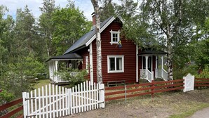 Une maison en bois rouge, dotée d’un porche blanc et d’une petite terrasse, entourée d’arbres et d’une clôture blanche en piquets.