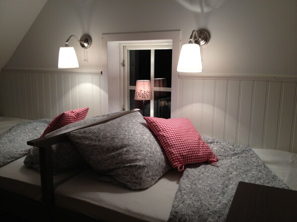 Un dormitorio con techo inclinado, dos lámparas de noche, una ventana con vistas y una cama con ropa de cama estampada y almohadas rojas.