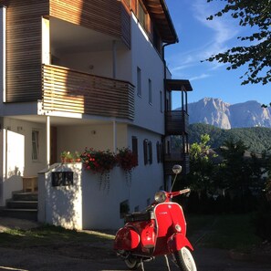 A two-story building with a balcony, a red vintage Vespa parked in front, and a mountainous backdrop. - Apartment Hintertrojer in a central location, good food with free WIFI (Lajen)
