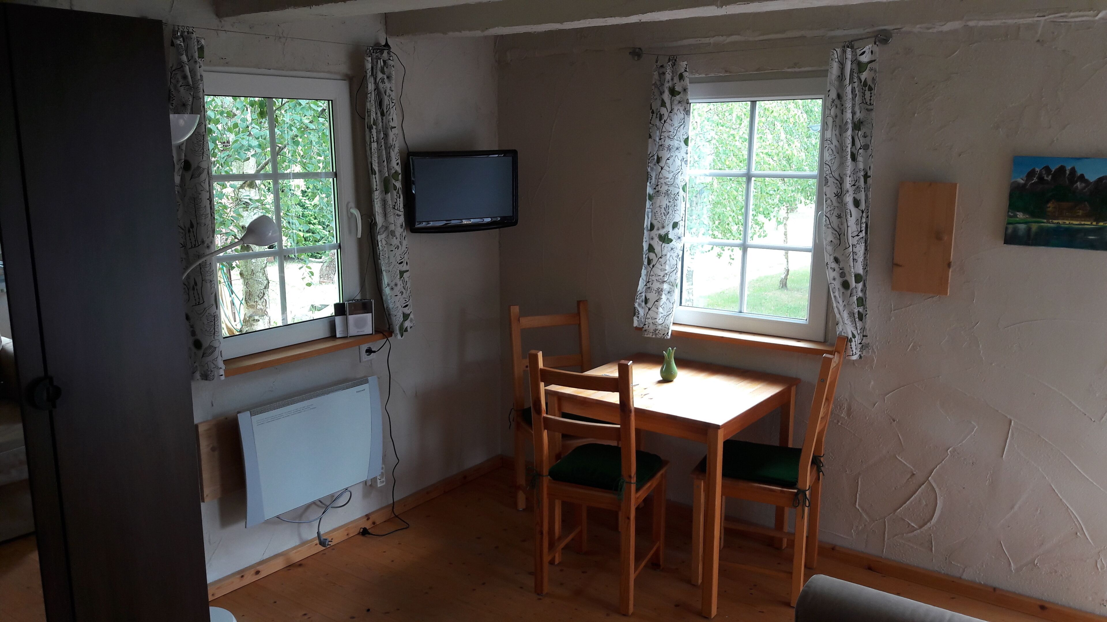 Ein Zimmer mit einem Holztisch, Stühlen, einem an der Wand befestigten Fernseher und zwei mit Vorhängen versehenen Fenstern.
