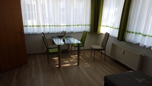 A room with a glass-top dining table, four chairs, a radiator, and a window with sheer curtains.