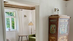 Et værelse med trægulv, en grøn lænestol, et højt skab med kunst, et bord og et vindue med gardiner.