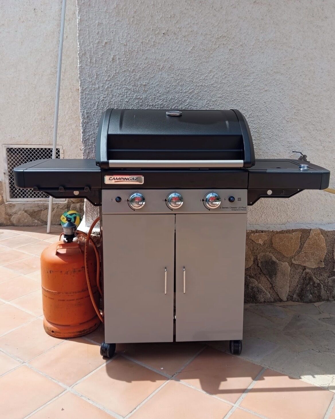Ein tragbarer Gasgrill mit schwarzem Deckel, drei Reglern und einer Propangasflasche.