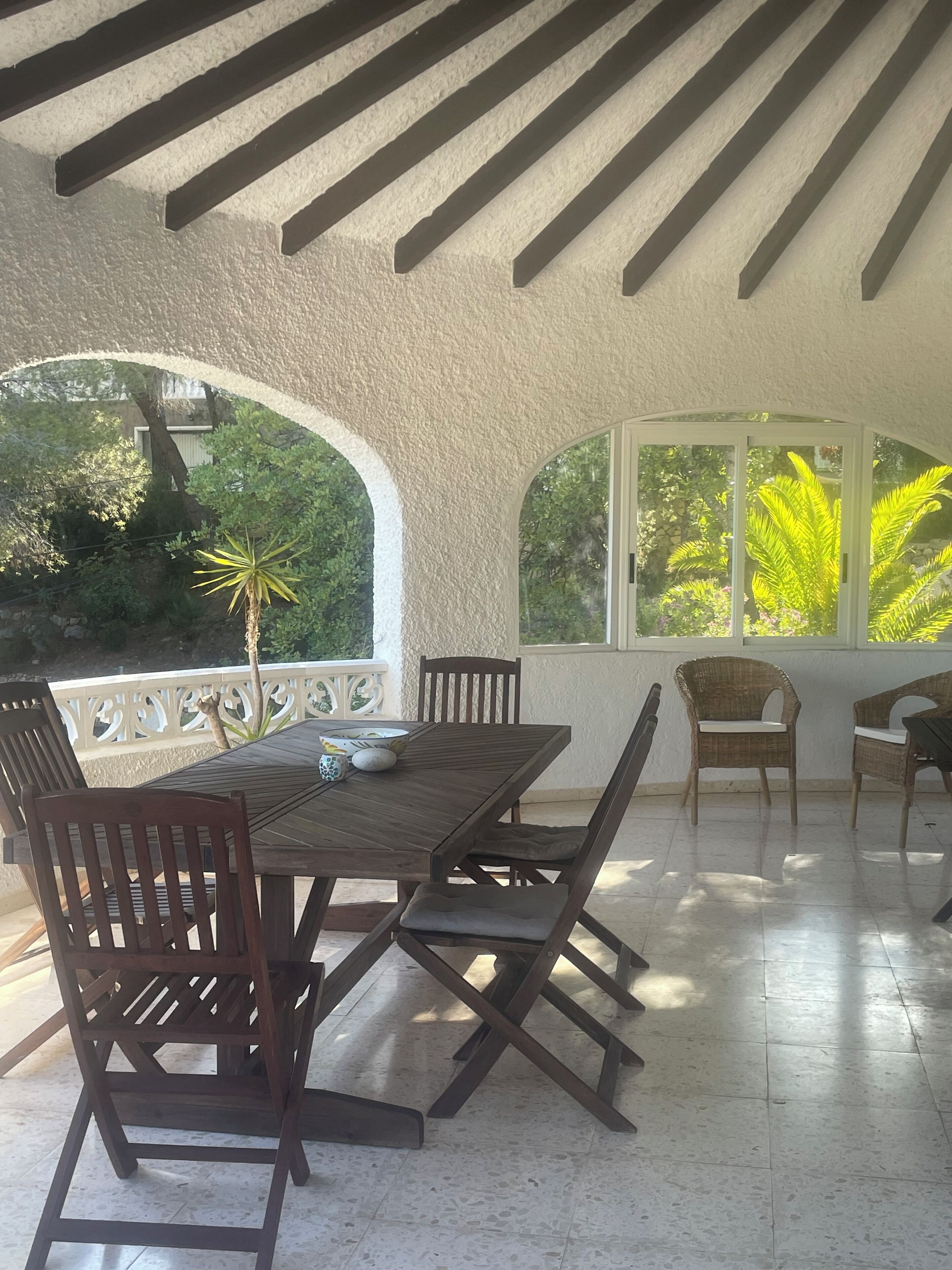 Un porche cubierto con mesa y sillas de madera, amplias ventanas y vista a la vegetación.