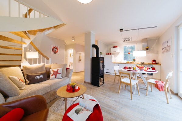 A modern living room with a staircase, a dining area with a table and chairs, and a kitchen with white cabinets and a black refrigerator.