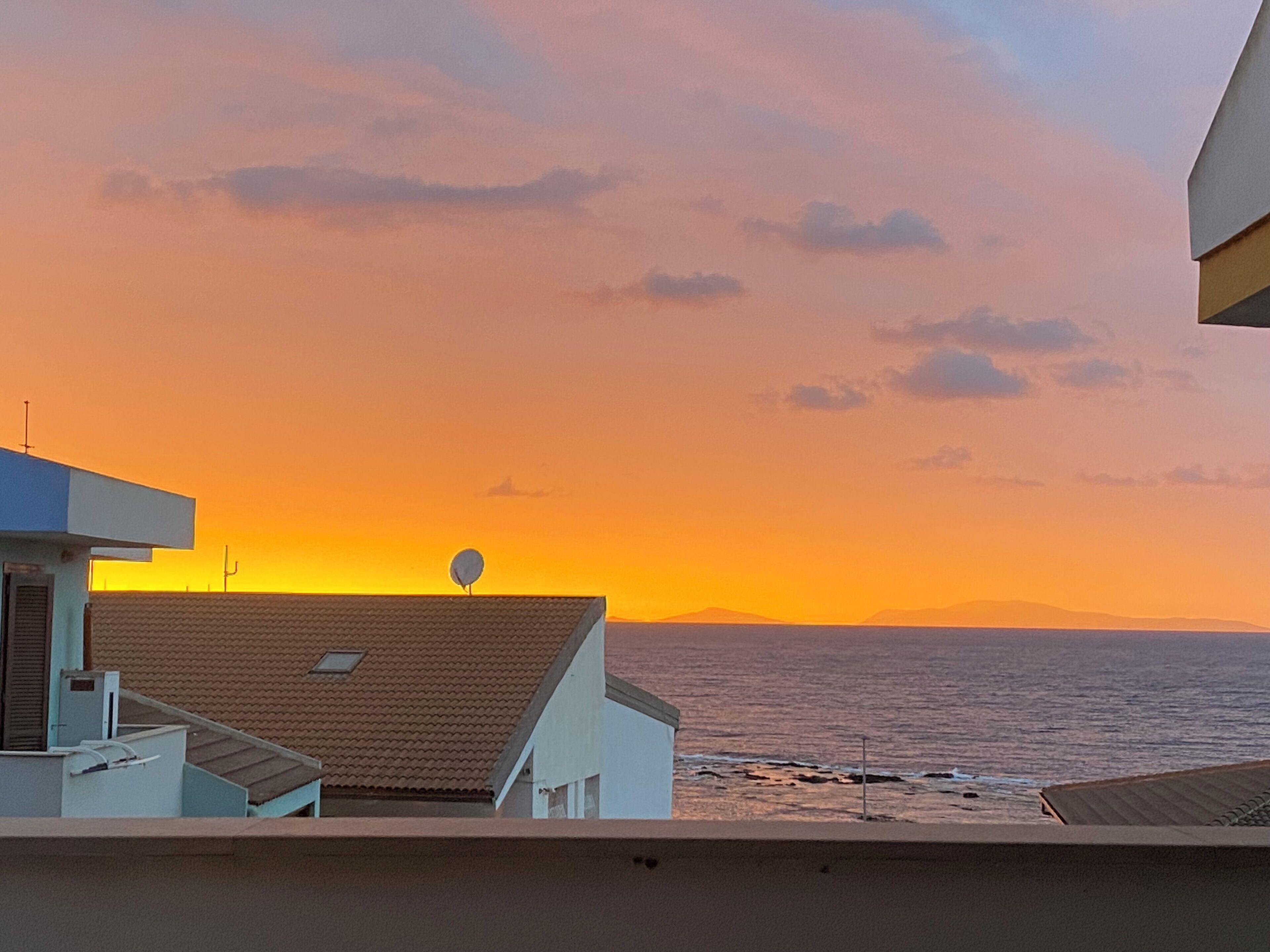 A coastal sunset view from a balcony overlooking a beach, with buildings in the foreground.