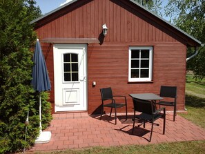 A red wooden building with a white door, two windows, and a patio with a table and chairs.