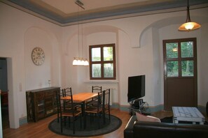 A dining area with a wooden table and chairs, a clock on the wall, a television, and a window with a view of greenery.