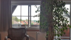 A room with a wicker chair, a large window with a view of houses, and a potted plant. - Top apartment in small complex, 2-4 pers. 65sqm, balcony, 2 bedrooms. W-lan (Walkenried)