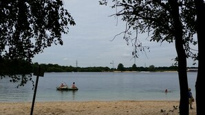 Una playa con un lago tranquilo, un bote pequeño con dos personas y un niño jugando en el agua.