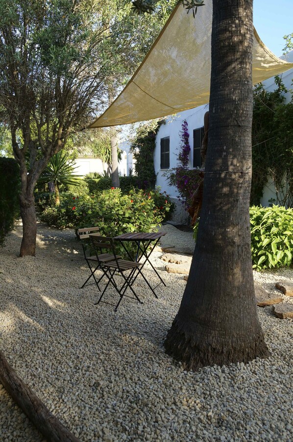 A garden with a table and chairs under a shade cloth, surrounded by gravel and trees.