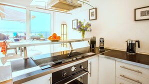 A modern kitchen with a gas stove, oven, and coffee maker. There is a window with a view of the sea, and a vase with flowers on the counter.