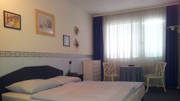 Quarto de hotel com cama, escrivaninha, duas cadeiras, cômoda e janela com cortinas.