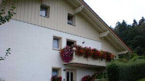 Un edificio de dos pisos con fachada blanca, un balcón con macetas de flores y un camino de piedra que conduce a la entrada.