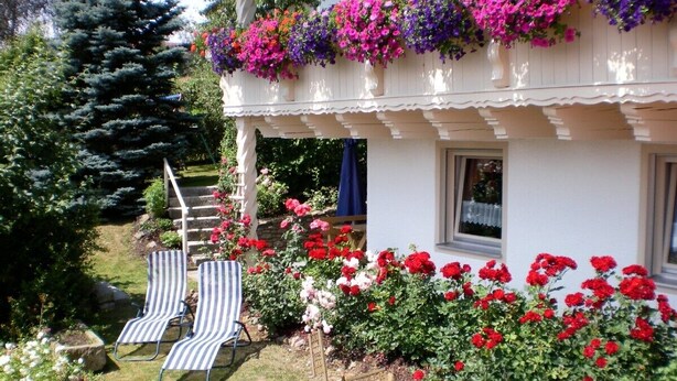 Una casa con balcón decorado con flores de colores, un pequeño jardín con flores rojas y blancas, y dos sillones de jardín a rayas.
