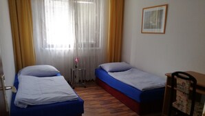 A room with two single beds, a small table, a chair, and a window with curtains.