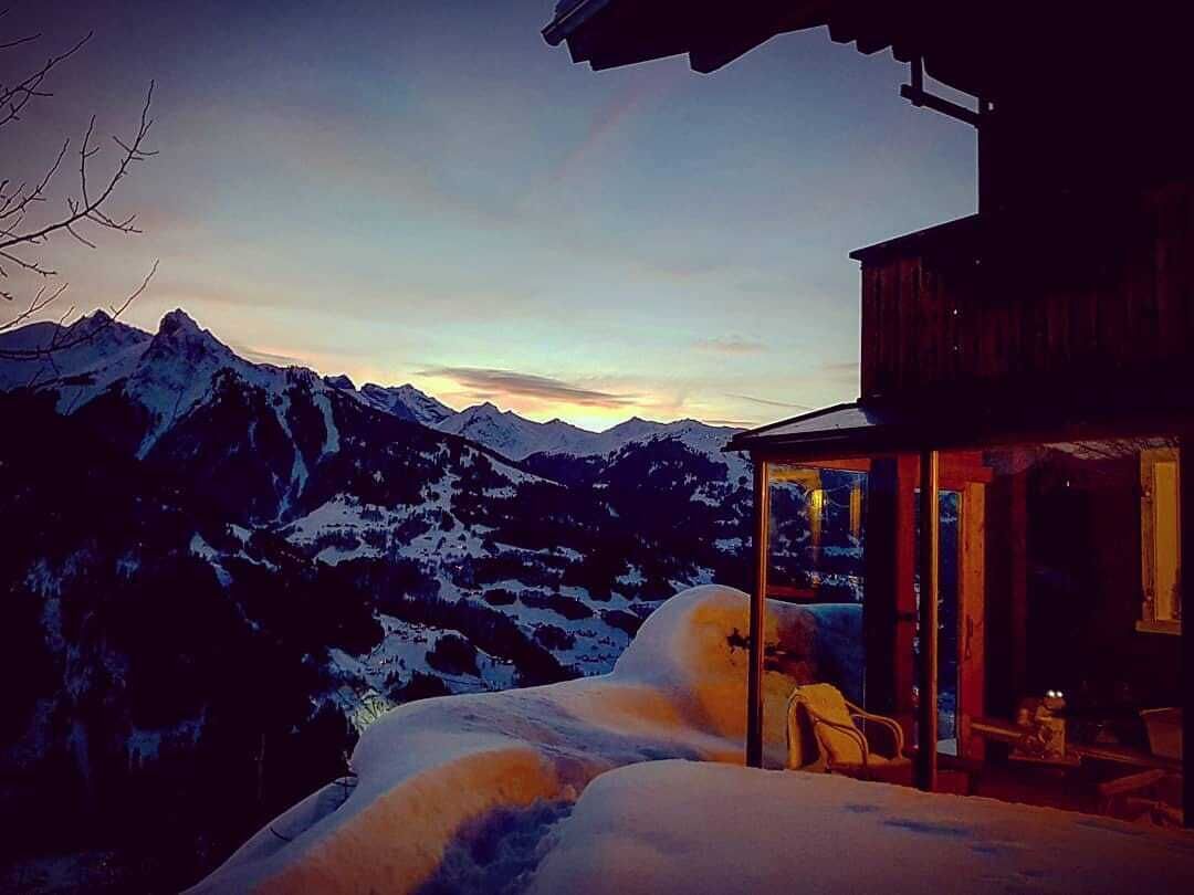 A mountain cabin with a glass door leading to a cozy interior, surrounded by snow-covered peaks.