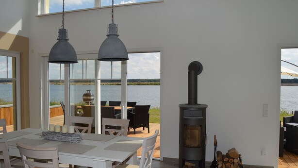 Un interior moderno con una gran mesa de comedor, dos lámparas colgantes, un estufa de leña y vistas al agua.