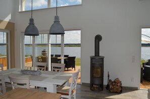 A modern interior with a large dining table, two pendant lights, a wood-burning stove, and a view of the water outside. - Private beach! NEW villa "Cloudless" with fireplace and sauna right on the beach (Löbnitz)