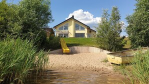 Una casa con un amplio ventanal, un espacio exterior cubierto y una zona arenosa con un bote.