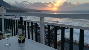 A balcony with a table, two wine glasses, and a bottle of wine. The view includes a sunset over the ocean and a distant town. - Apartment directly on the sea with a fantastic view in Punta del Hidalgo (La Laguna / Punta del Hidalgo)