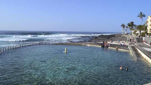 A clear pool with a curved edge, a rocky shoreline, and a beach with palm trees and people in the background. - Apartment directly on the sea with a fantastic view in Punta del Hidalgo (La Laguna / Punta del Hidalgo)