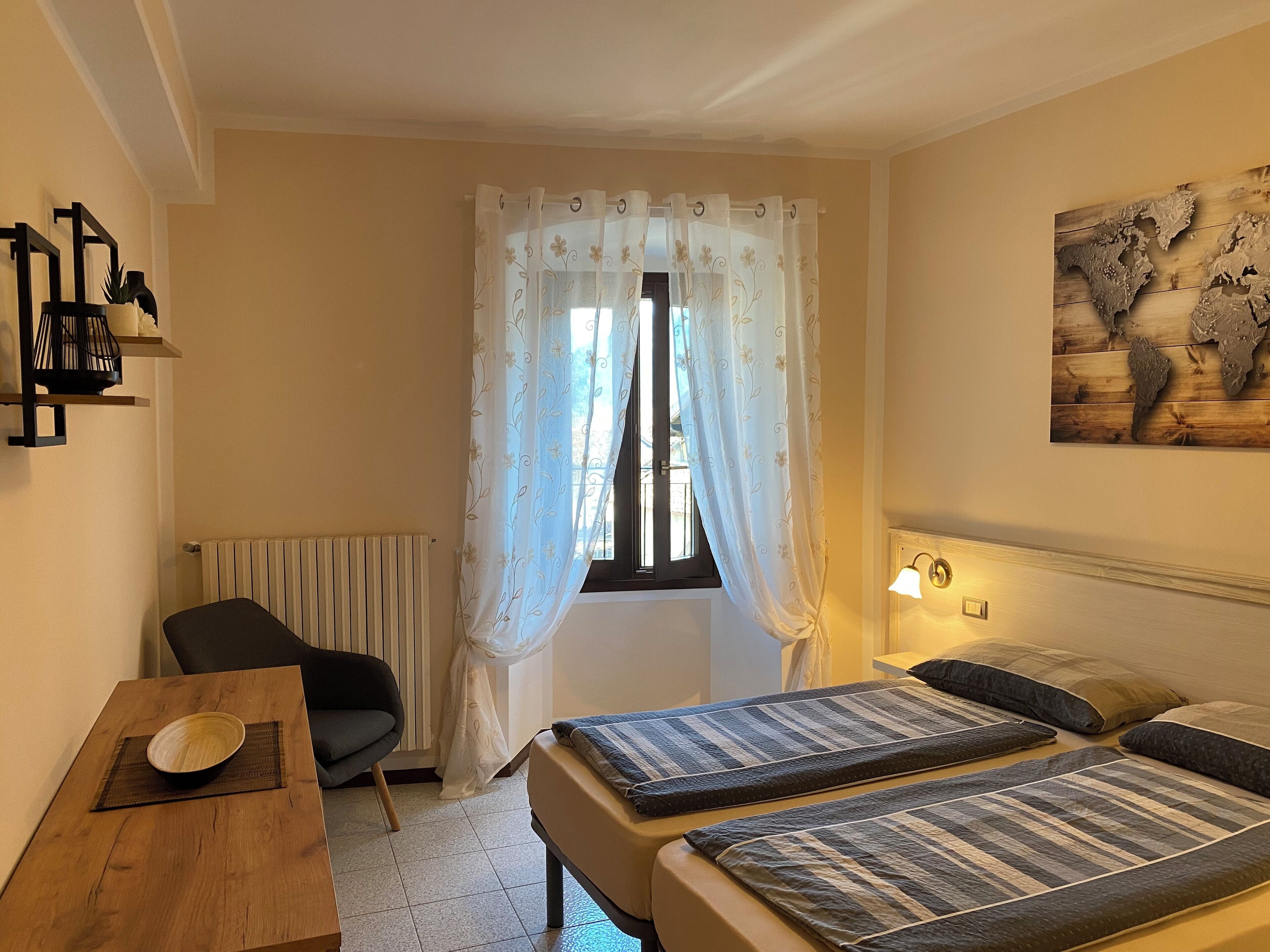 Hotellihuone, jossa on sänky, tuoli, pöytä ja kulho, ikkuna verhoineen, patteri ja seinään kiinnitetty lamppu.