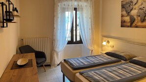 Et hotelværelse med seng, stol, bord med skål, vindue med gardiner, en radiator og en vægmonteret lampe.