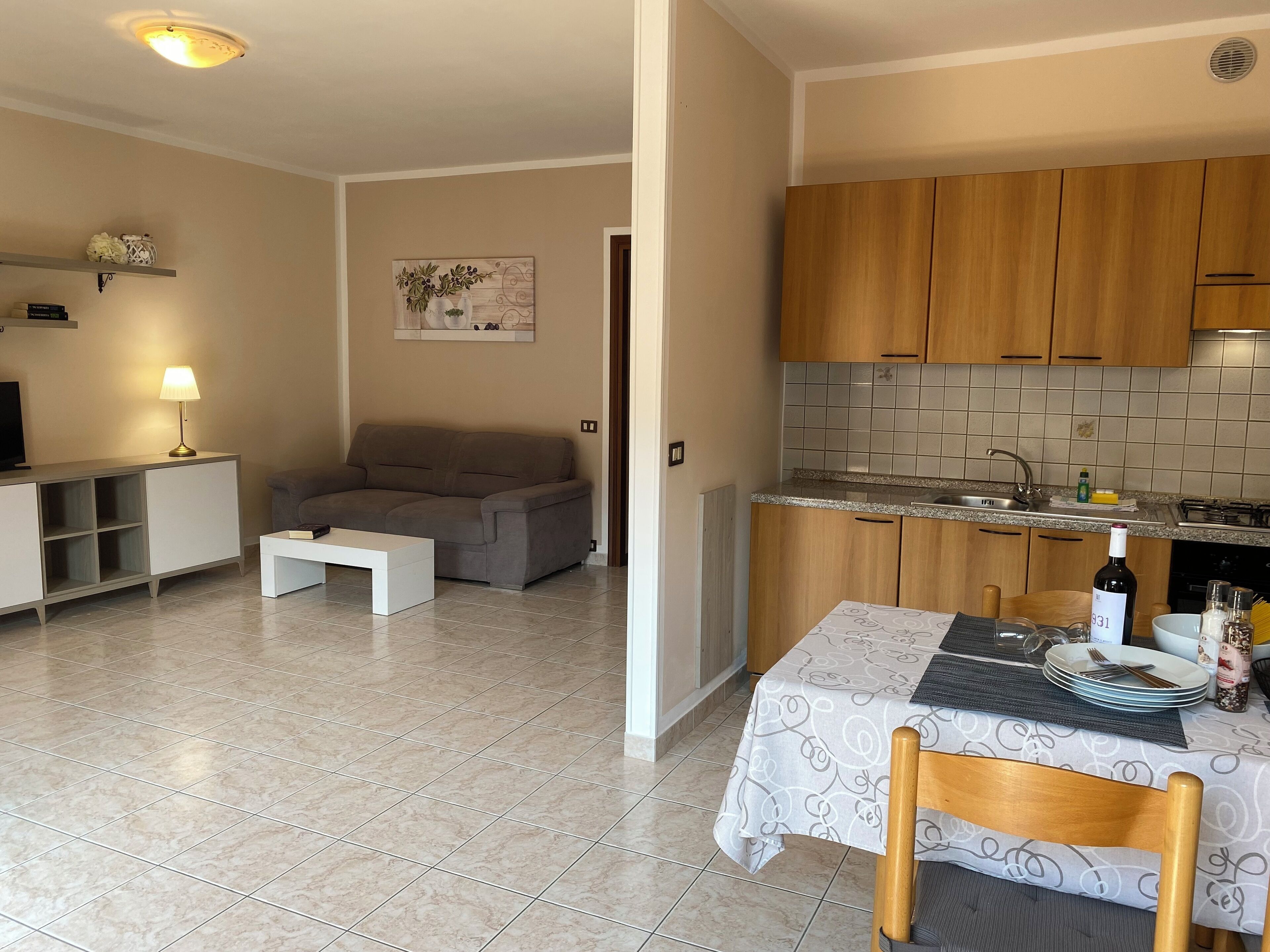 A living room with a sofa, coffee table, and a TV. There's a dining area with a table set for two, a kitchen with wooden cabinets, and a tiled backsplash.