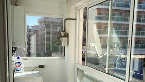 A small balcony with a white chair, a table with a bottle of detergent, and a view of buildings through a large window.