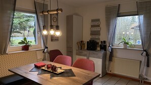 Un comedor con mesa de madera, sillas rojas y una encimera de cocina con cafetera. Hay plantas en macetas en el alféizar de la ventana y un reloj de pared.