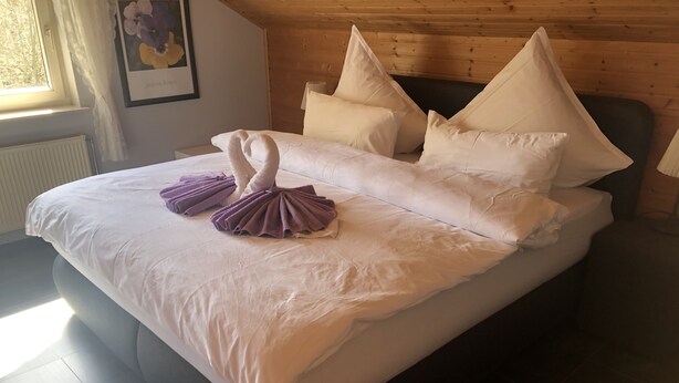 Una cama impecablemente arreglada con un diseño de cisne en la toalla, una obra de arte floral enmarcada en la pared y un cabecero de madera.