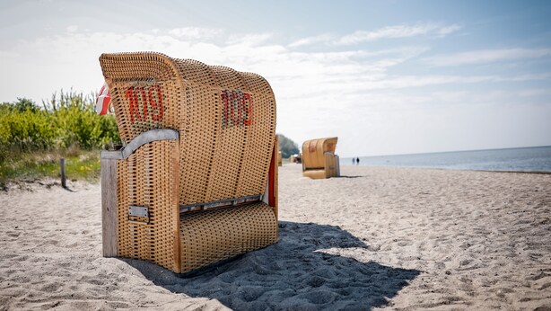 A wicker beach chair with a metal frame and a cushion, placed on a sandy beach with a clear sky and ocean in the background.