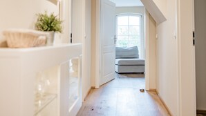 A narrow hallway with a wooden floor leading to a room with a window and a grey sofa.