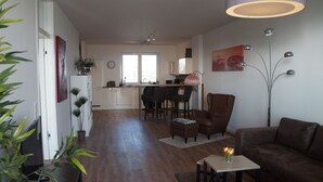 A modern living room with a brown leather sofa, a small coffee table, and a TV on a stand. There is a dining area with a table and chairs in the background.