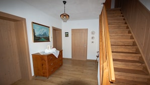 Una escalera de madera que lleva a un segundo piso, un aparador de madera con cajones, un cuadro de un paisaje montañoso y una puerta con una pequeña imagen encima.